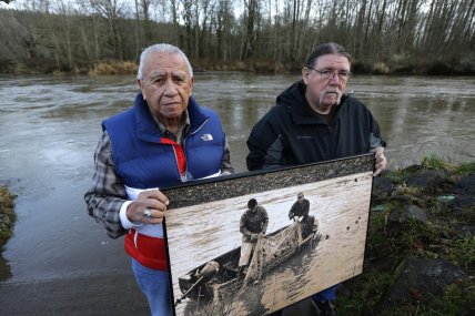 Billy Frank Jr., left, a Nisqually tribal elder who was arrested dozens of times while trying to assert his Native fishing rights during the Fish Wars of the 1960s and ’70s, poses for a photo Monday, Jan. 13, 2014, with Ed Johnstone, of the Quinault tribe, at Frank’s Landing on the Nisqually River in Nisqually, Wash. They are holding a photo from the late 1960s of Frank and Don McCloud fishing on the river. (AP Photo/Ted S. Warren)